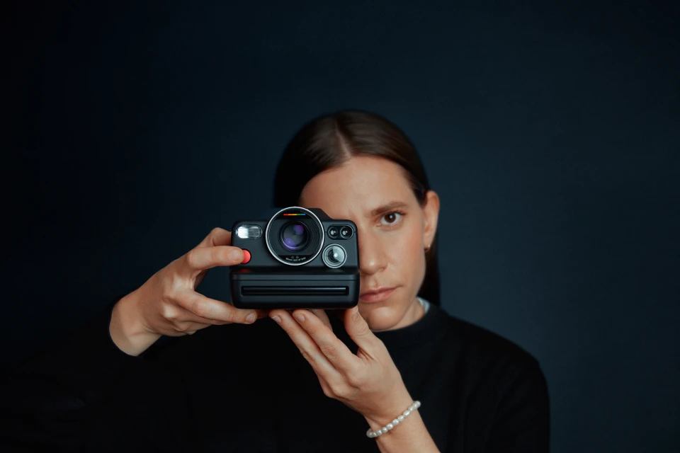 Adult female holding a black Polaroid camera against a dark blue background in a studio, capturing a modern and simple aesthetic.