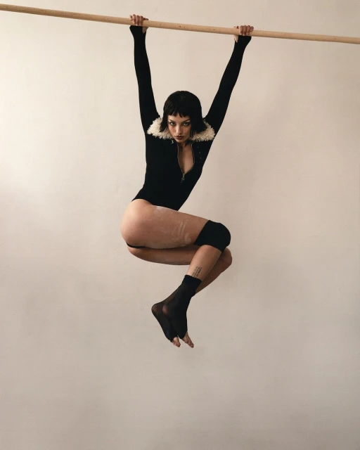 Athletic young woman in a black bodysuit with a white faux-fur collar suspended from a wooden bar in front of a plain white background.