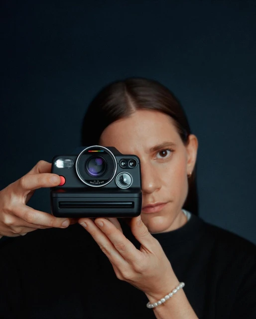 Adult female holds a black Polaroid camera in front of her face with both hands, eye looking over camera at viewer, dark background.