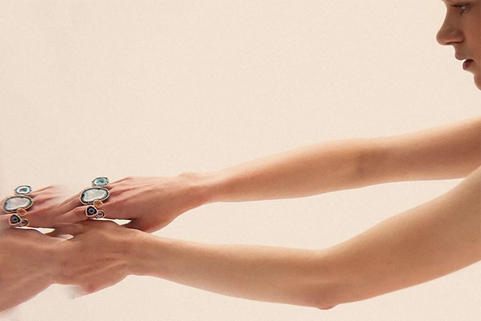Close-up of two adult females' arms overlapping, showcasing two fashion rings with gemstones, set against a pale background.
