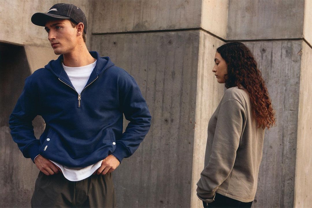 Two people wearing casual clothing stand in front of a concrete wall on an overcast day in an outdoor setting.