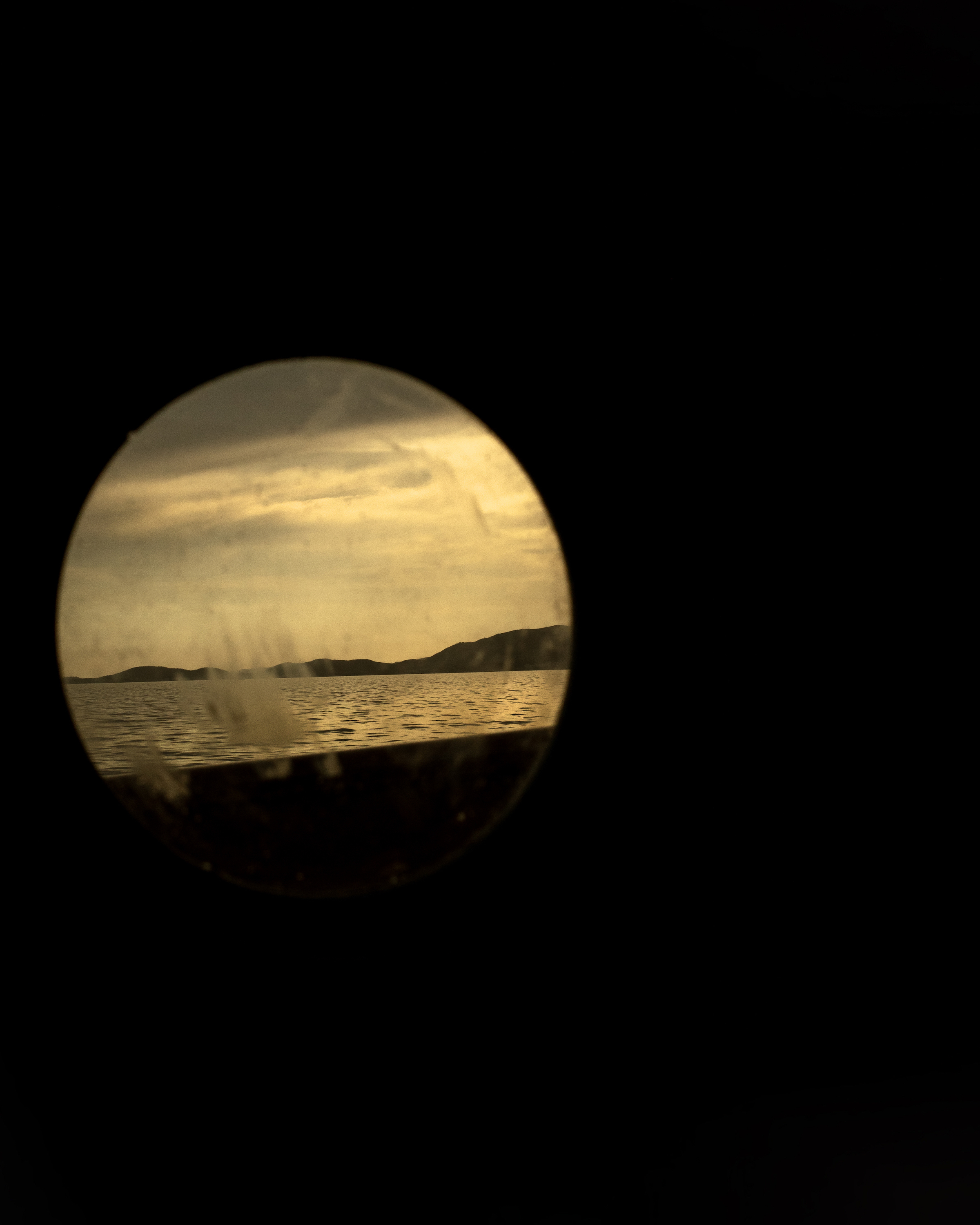 Scenic view through a circular frame: serene lake meeting distant hills under an overcast sky, shot with muted, sepia-toned color grading.
