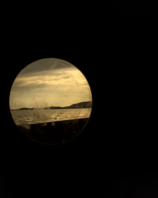 Scenic view through a circular frame: serene lake meeting distant hills under an overcast sky, shot with muted, sepia-toned color grading.