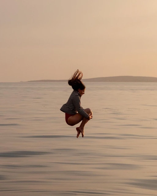 Woman jumps in the air over the ocean wearing a striped jacket and red bottoms with an island in the background on a sunny day.