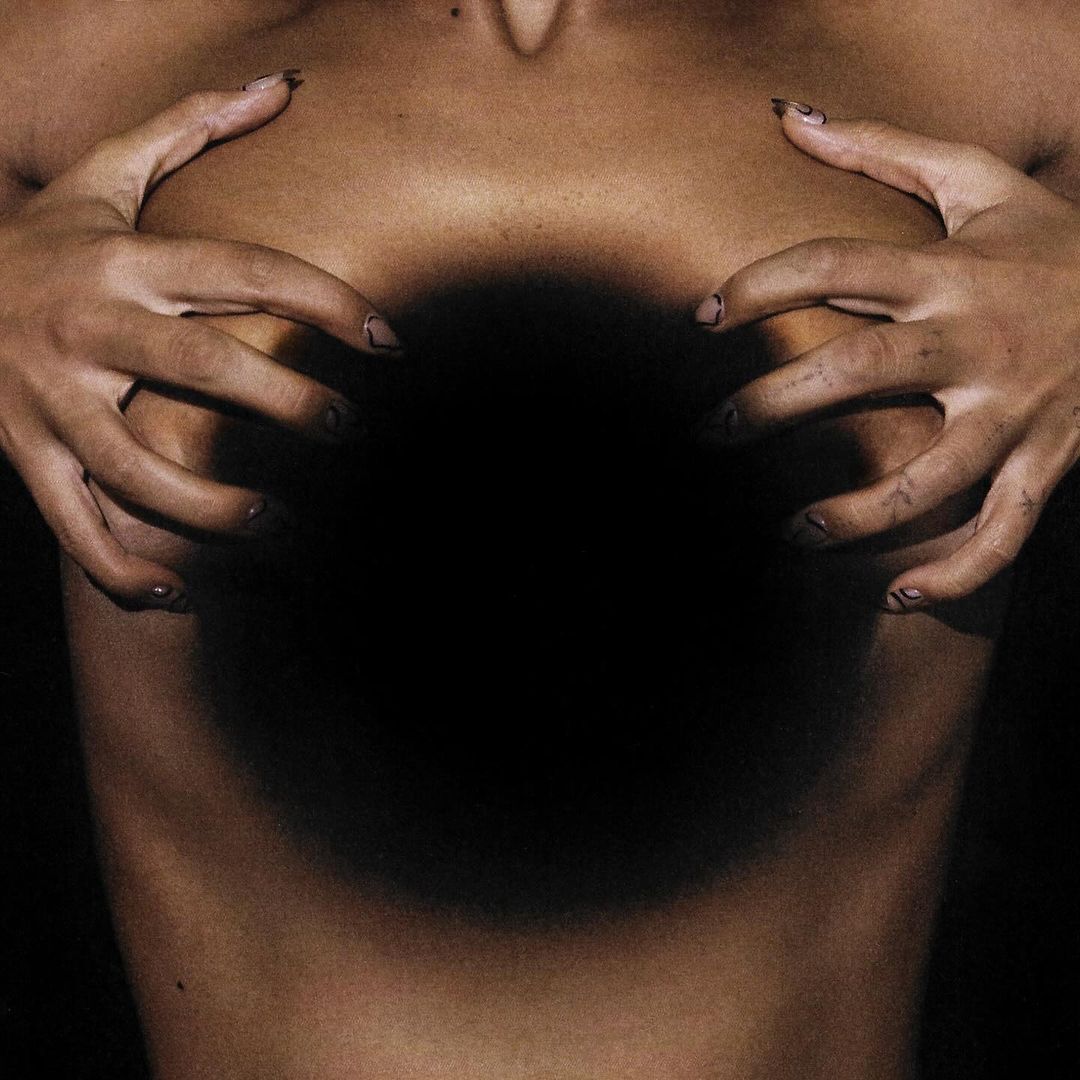 Close-up studio shot of a fair-skinned adult female holding her breasts, showcasing manicure details and dramatic lighting.