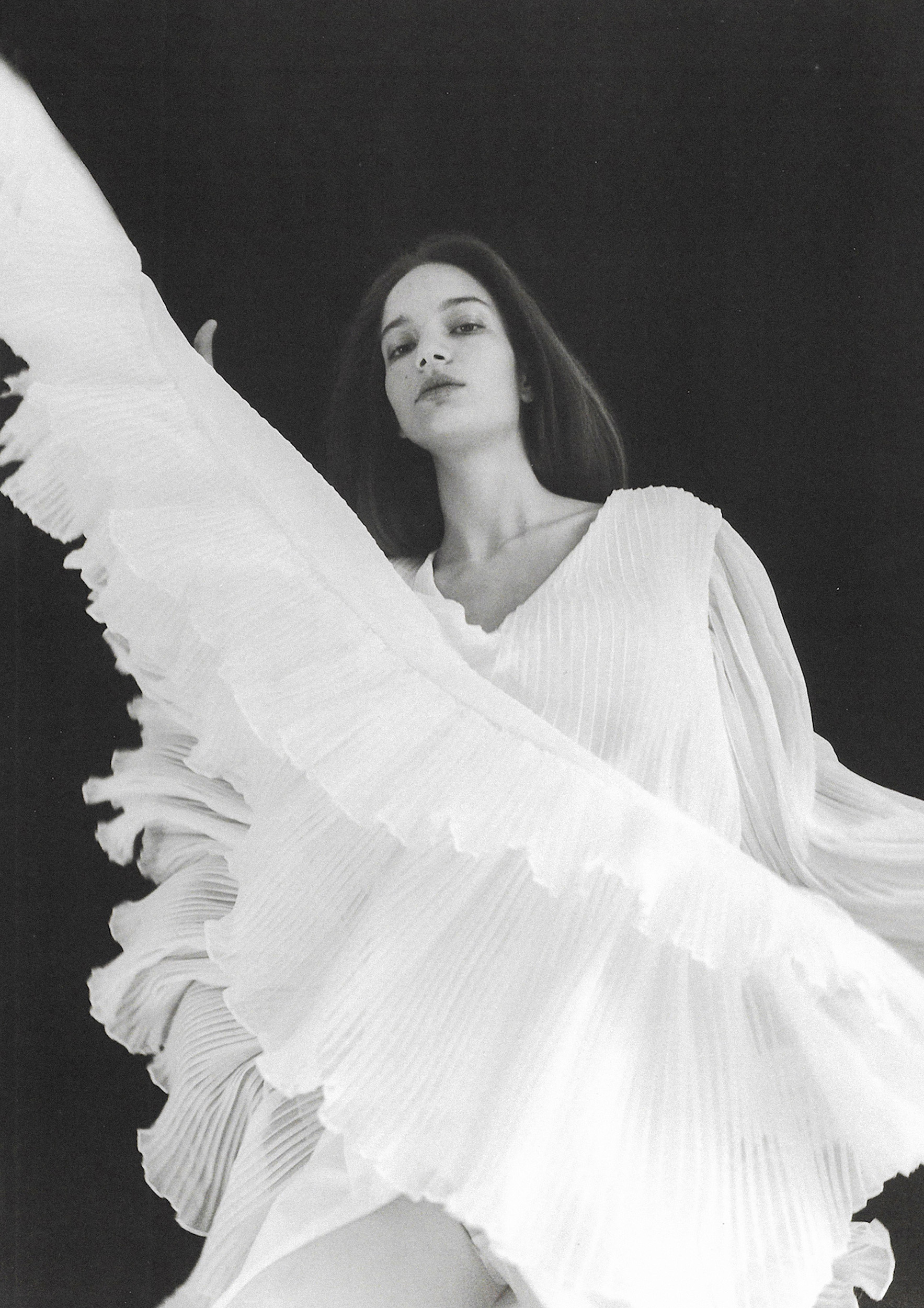 Monochrome portrait of a woman in a flowing pleated white dress captured in mid-motion against a dark background.