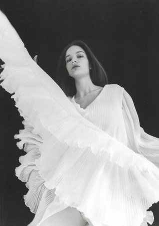 Monochrome portrait of a woman in a flowing pleated white dress captured in mid-motion against a dark background.