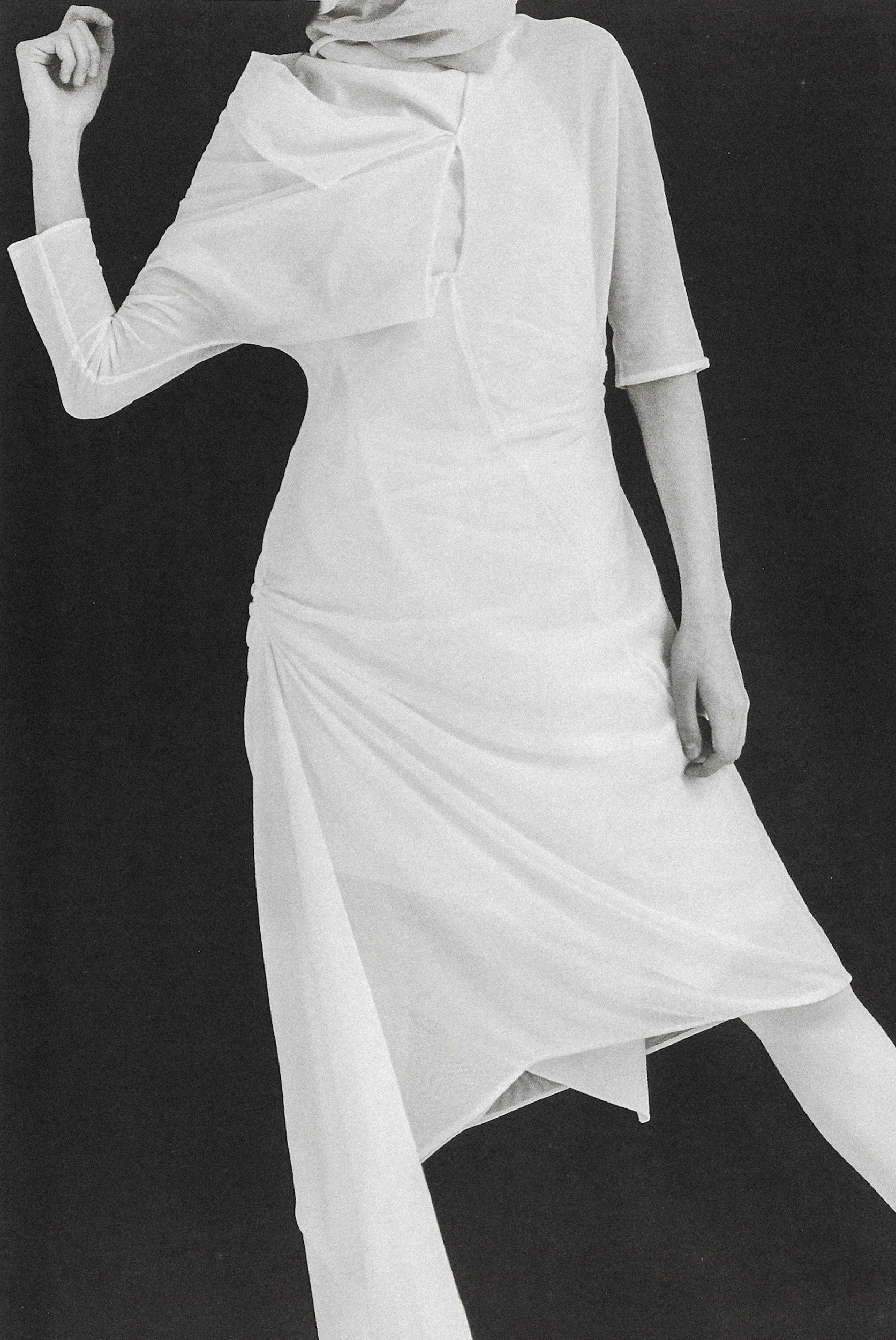 Fashion shoot featuring a slender woman in a flowing white dress with unique draping against a dark background in a studio environment.