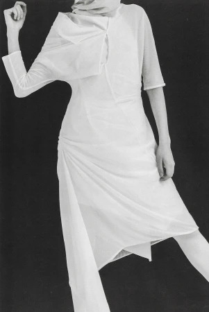 Fashion shoot featuring a slender woman in a flowing white dress with unique draping against a dark background in a studio environment.