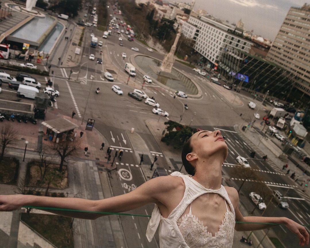Woman in elegant dress gazes upward in urban outdoor setting with distant cityscape and cars, evoking a sense of freedom and urban fashion.