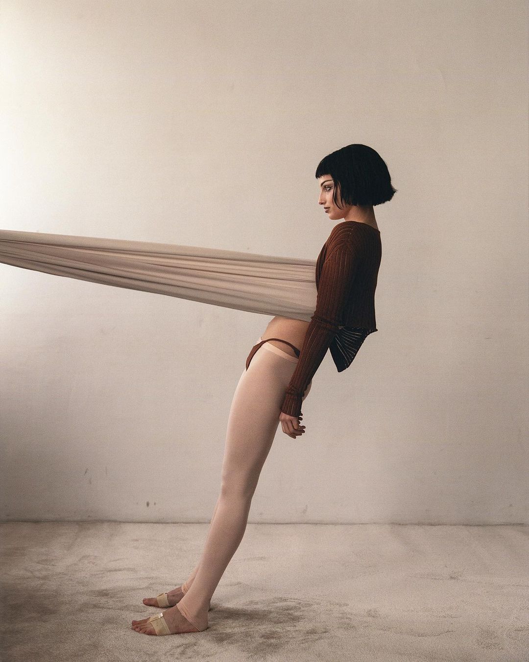 Woman in brown top and beige bottoms leans against a beige fabric in a minimalist studio setting, creating a contemporary fashion image.