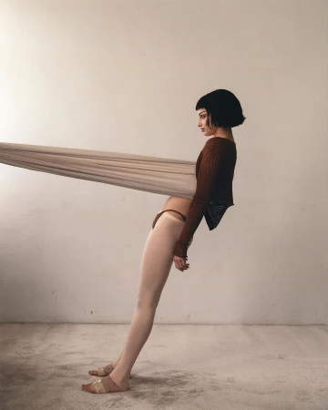 Woman in brown top and beige bottoms leans against a beige fabric in a minimalist studio setting, creating a contemporary fashion image.