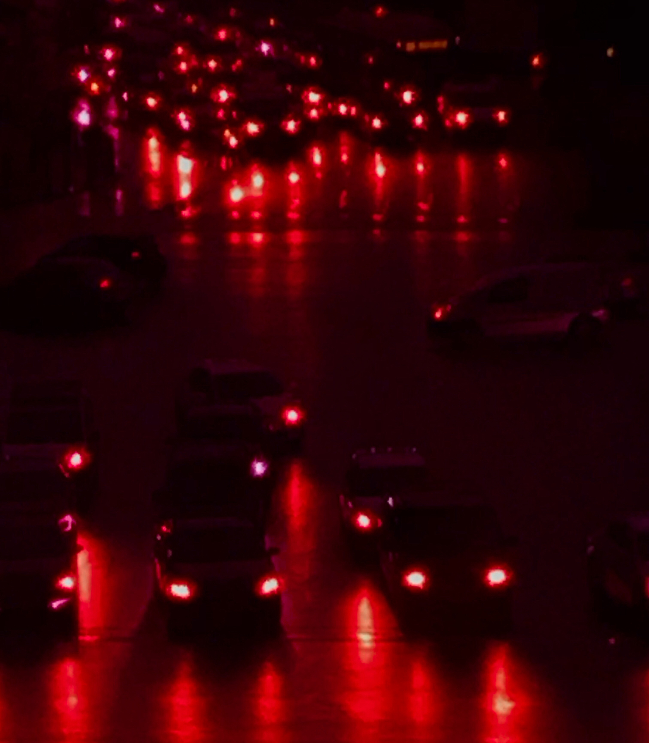 Night view of dark-colored automobiles on a city street, bright tail lights reflecting on the wet road surface, red color grading.