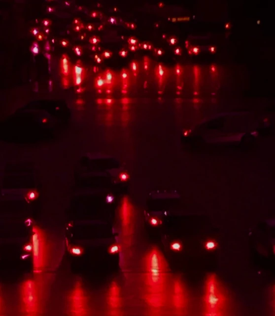 Night view of dark-colored automobiles on a city street, bright tail lights reflecting on the wet road surface, red color grading.