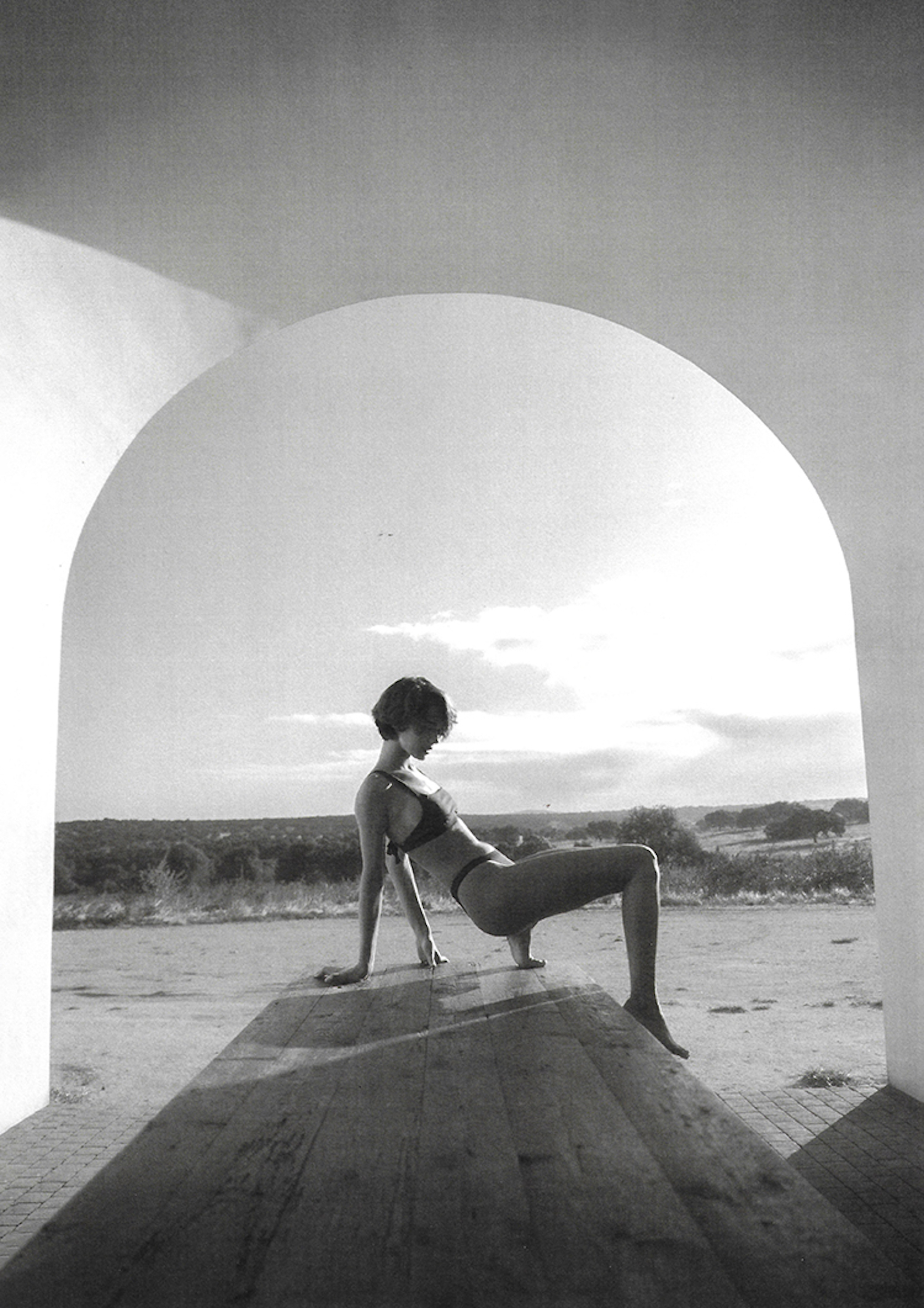 Monochrome portrait of a woman in a bikini top and bottoms posing on a wooden surface, shot through an arched opening outdoors.