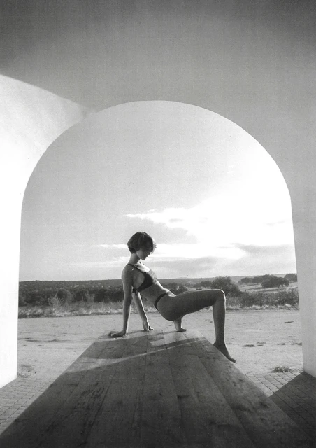 Monochrome portrait of a woman in a bikini top and bottoms posing on a wooden surface, shot through an arched opening outdoors.