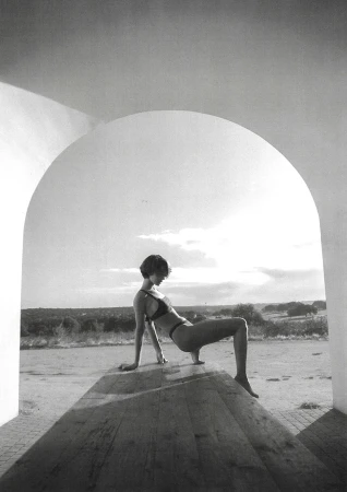 Monochrome portrait of a woman in a bikini top and bottoms posing on a wooden surface, shot through an arched opening outdoors.
