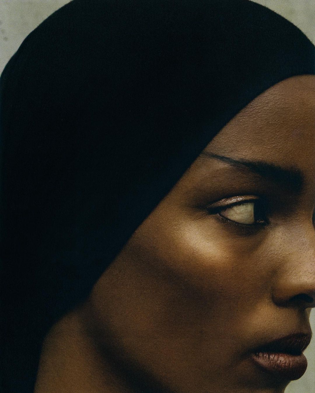 Close-up portrait of a dark-skinned woman wearing a dark headscarf, facing right, featuring gold eyeshadow and visible facial skin texture.