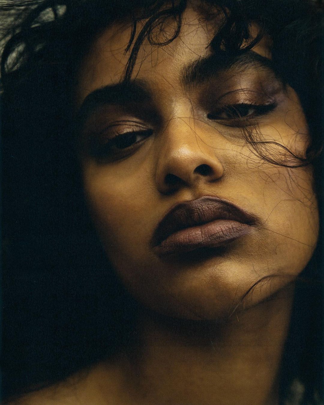 Close-up portrait of a woman with curly hair and dark lipstick against a dark background, emphasizing her facial features and skin tone.