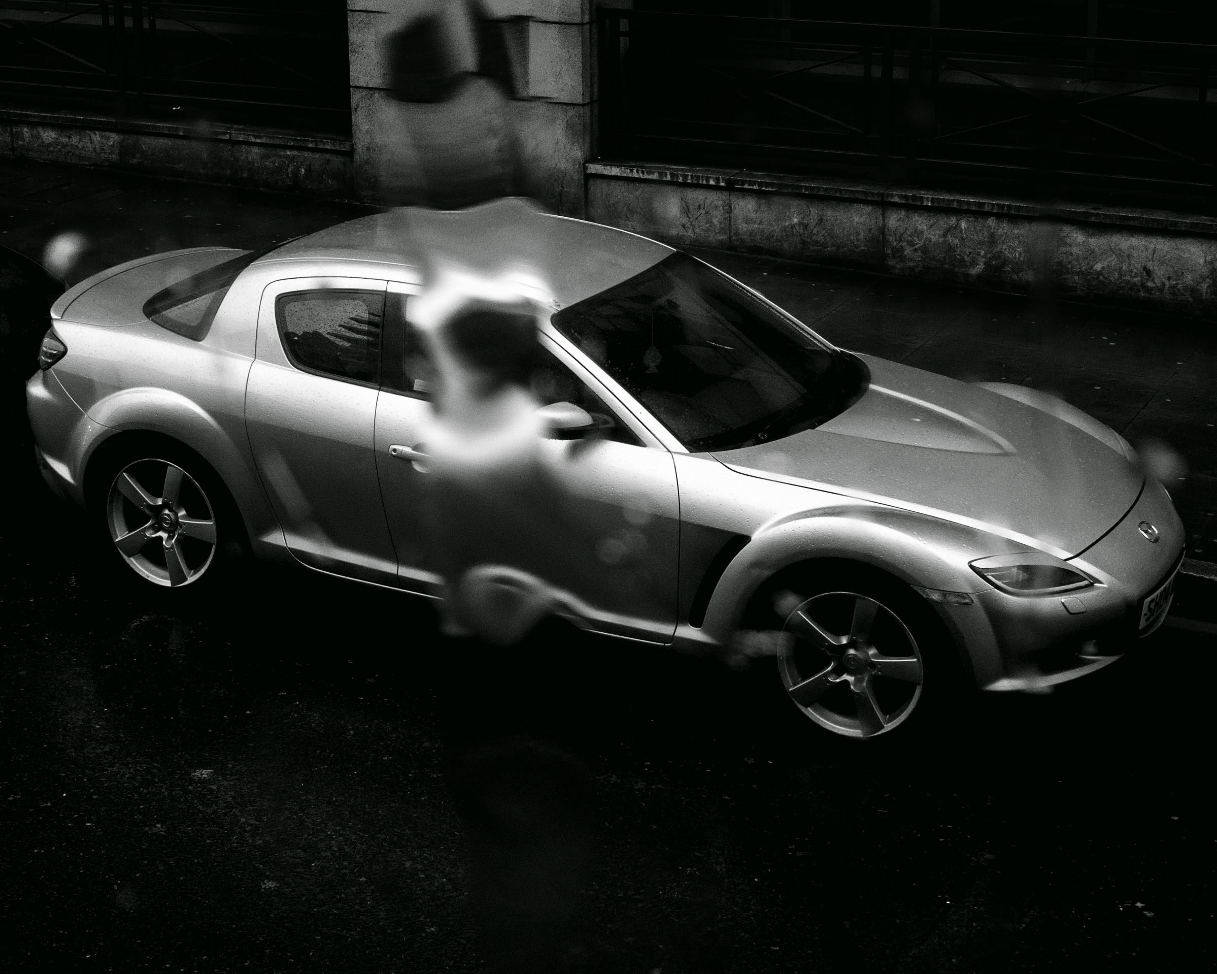 Monochromatic shot of a silver Mazda RX-8 sports car on dark asphalt, with a blurred figure in the background, creating a moody automotive image.