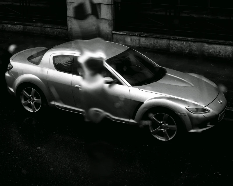 Monochromatic shot of a silver Mazda RX-8 sports car on dark asphalt, with a blurred figure in the background, creating a moody automotive image.