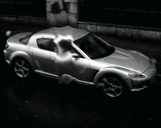 Monochromatic shot of a silver Mazda RX-8 sports car on dark asphalt, with a blurred figure in the background, creating a moody automotive image.