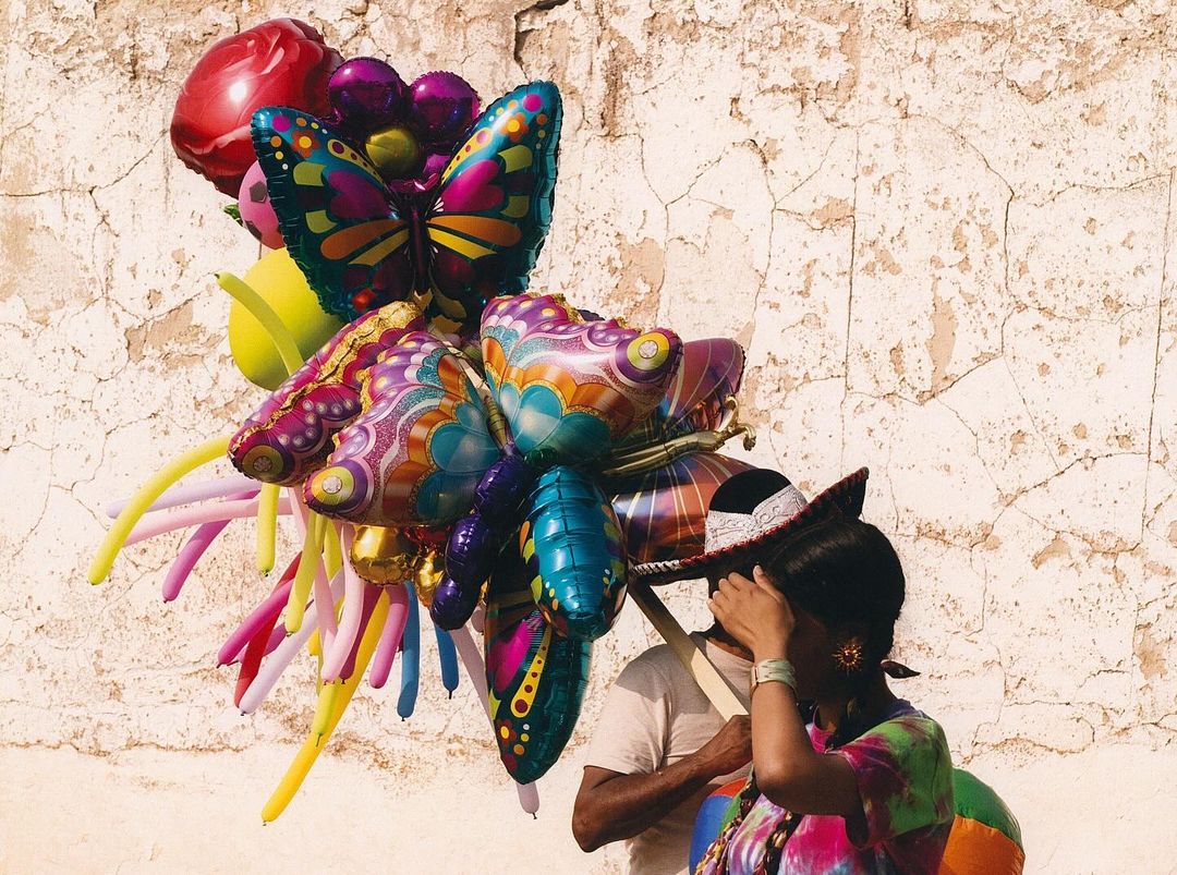 Woman carrying colorful butterfly and heart-shaped balloons in an outdoor setting with textured wall background.