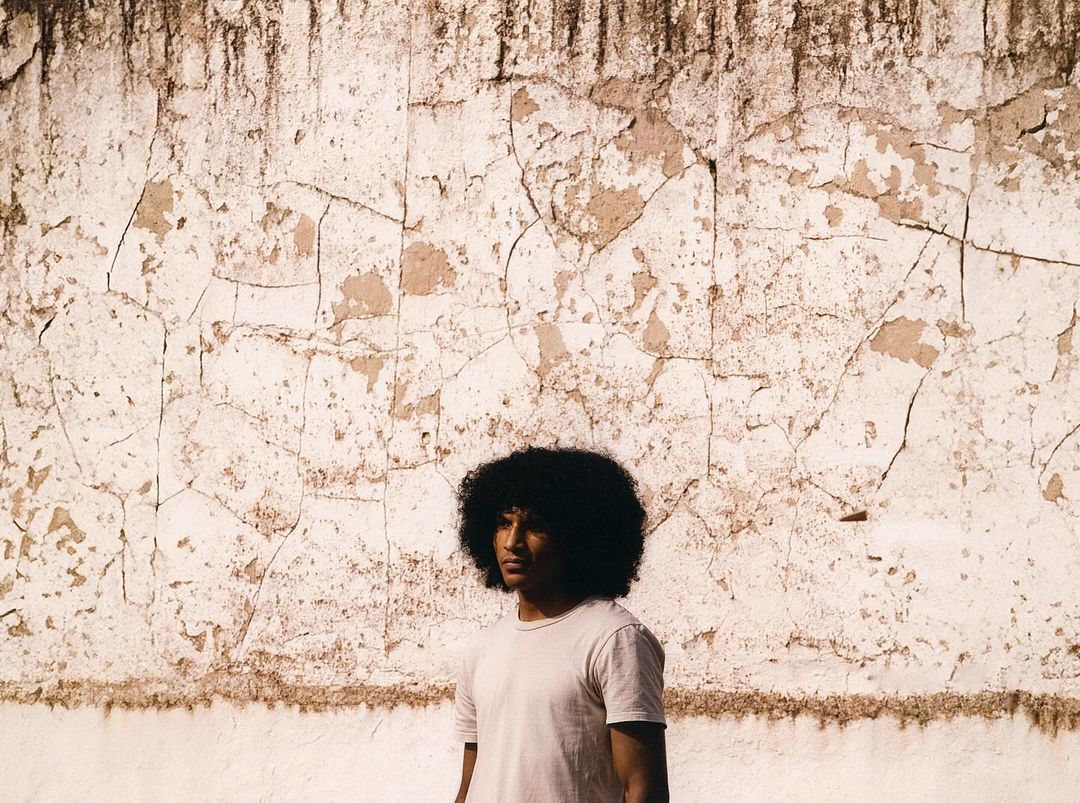 Adult male portrait in front of a weathered wall. The man, wearing a t-shirt, looks off to the side in outdoor daylight.