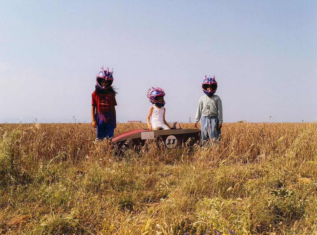 Three children wearing racecar driver helmets stand next to a box car in a field of tall golden wheat under a clear sky.
