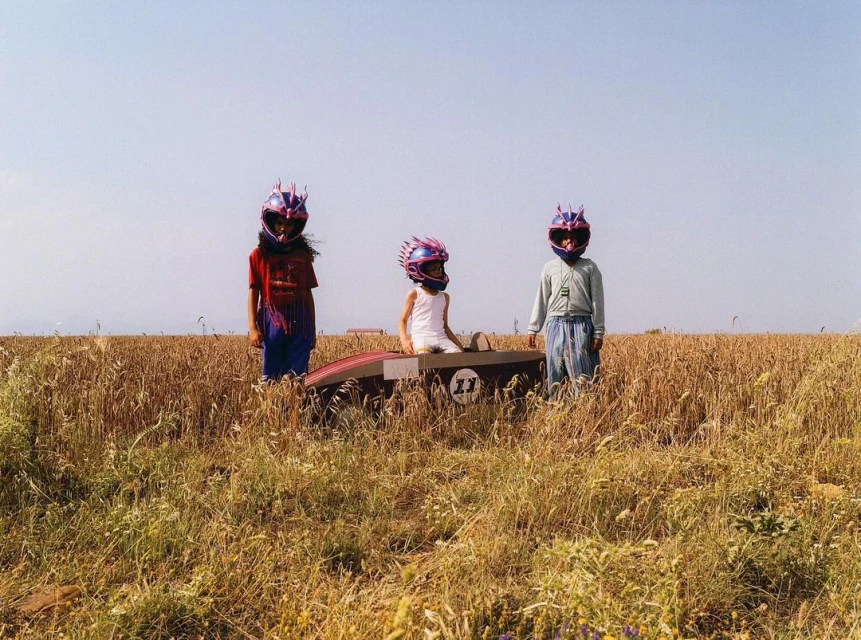 Three children wearing racecar driver helmets stand next to a box car in a field of tall golden wheat under a clear sky.