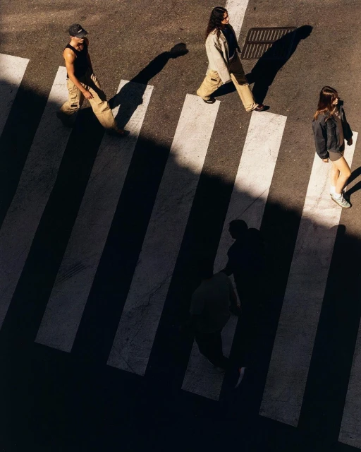 Three pedestrians walk across a zebra crossing on a sunny day, casting long shadows. Aerial view with bold black and white contrast.