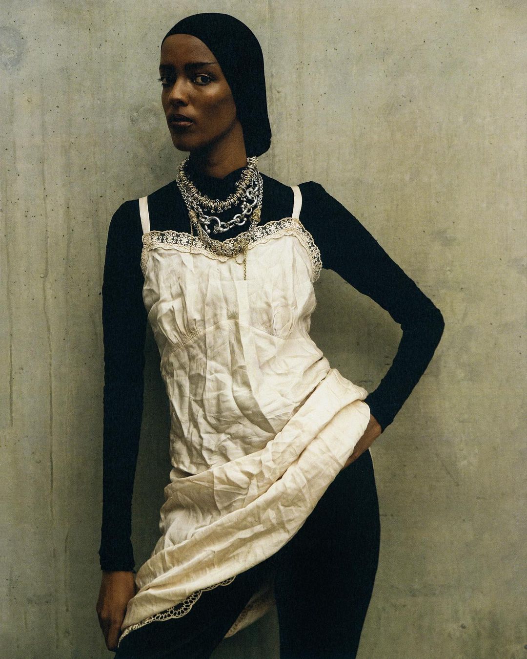 Woman in a black headscarf, top, and leggings stands against a gray wall, accessorized with multiple silver chain necklaces.