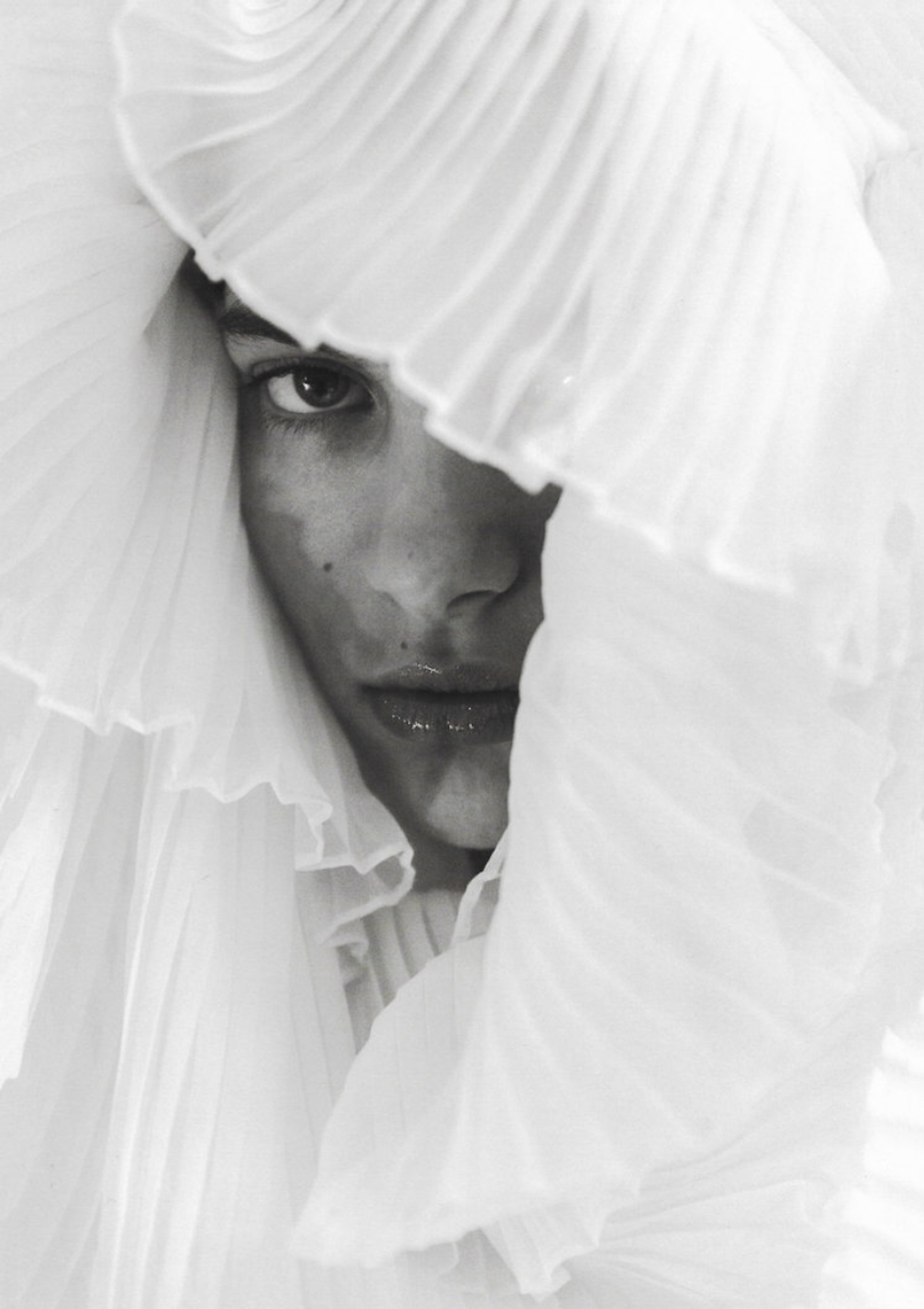 Close-up of a woman's face partially obscured by white ruffled fabric, creating an intimate and artistic fashion portrait with soft lighting.