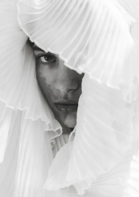 Close-up of a woman's face partially obscured by white ruffled fabric, creating an intimate and artistic fashion portrait with soft lighting.