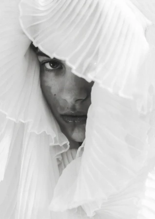 Close-up of a woman's face partially obscured by white ruffled fabric, creating an intimate and artistic fashion portrait with soft lighting.