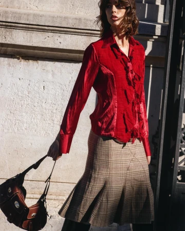 Model in red satin blouse and plaid skirt holding a brown leather handbag outdoors by stone building
