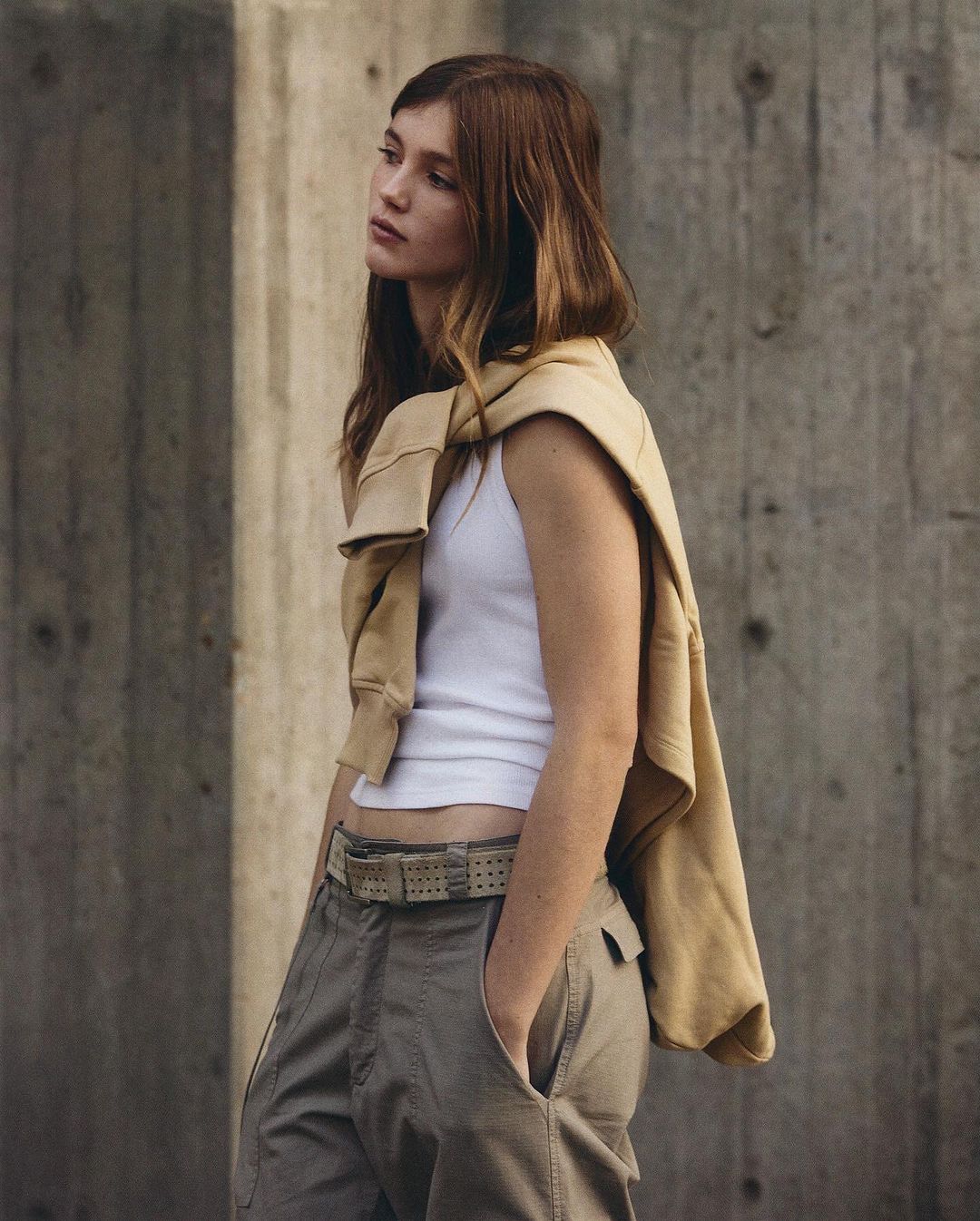 Woman in a white tank top and tan cargo pants stands against a concrete wall with a sweatshirt draped over her shoulders.