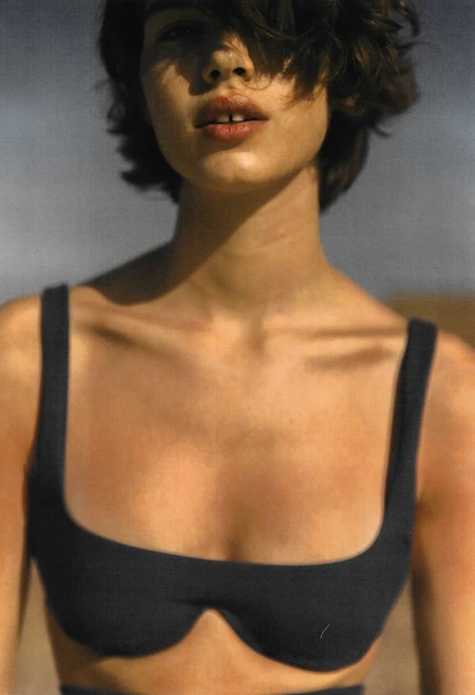 Close-up portrait of a brunette adult woman in a black bra top, with short hair, against a muted, neutral, outdoor background.