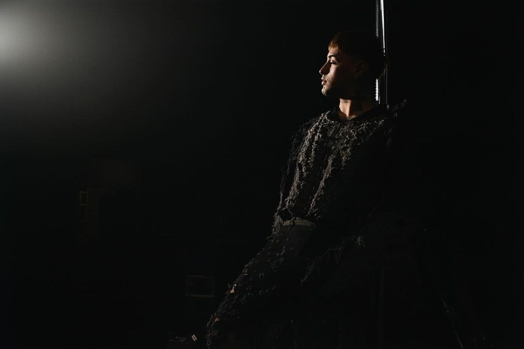 Portrait of a young adult male in textured dark clothing looking left in a dimly lit indoor space with dark background and spotlight.