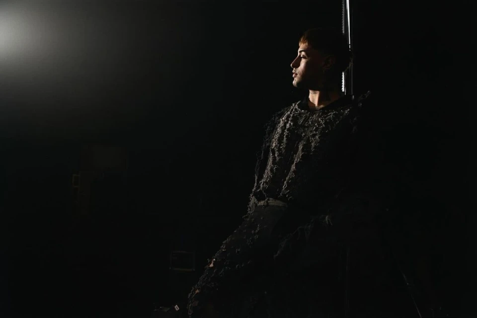 Portrait of a young adult male in textured dark clothing looking left in a dimly lit indoor space with dark background and spotlight.