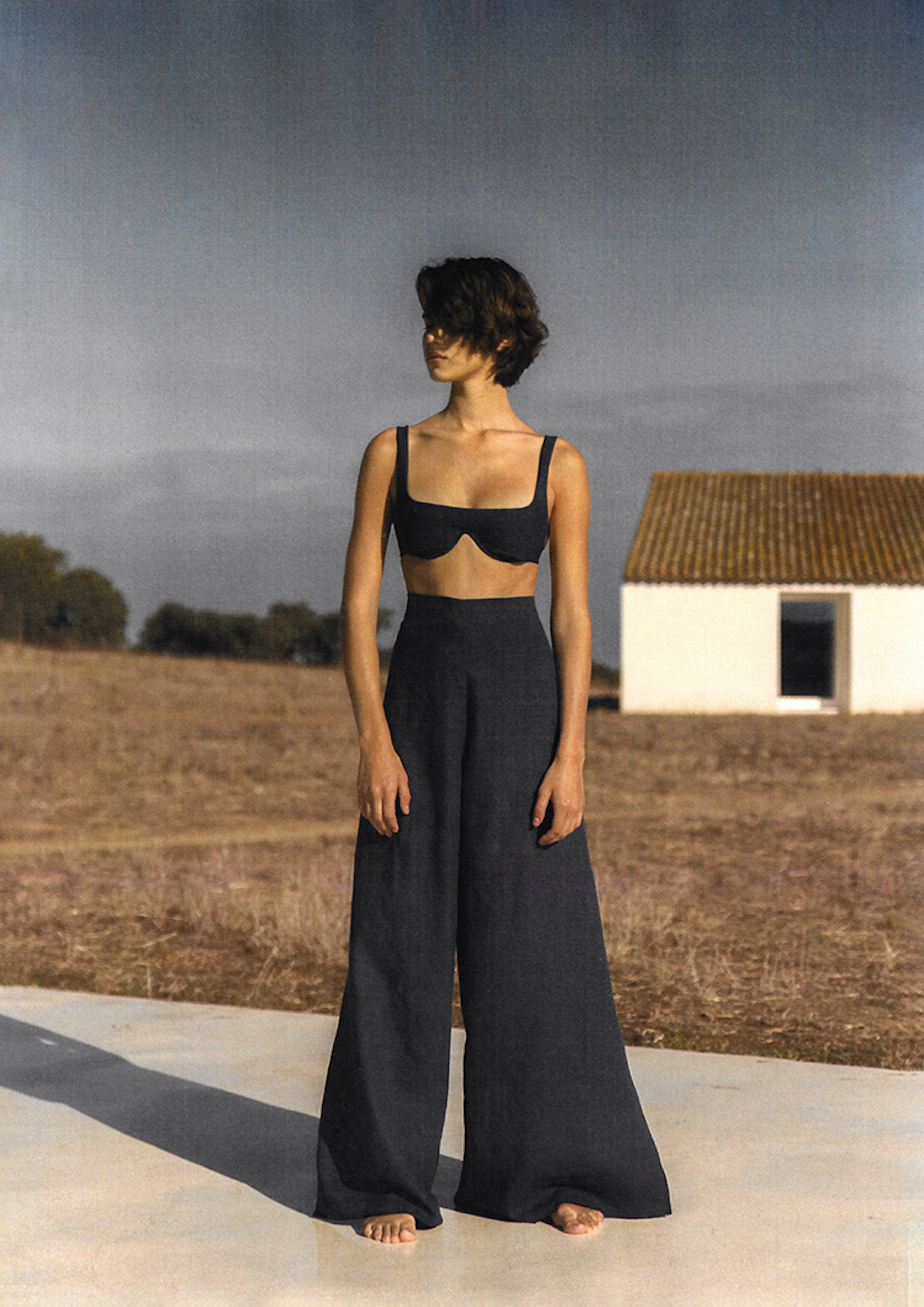 A woman wearing a black bikini top and loose black pants poses in front of a field and small white house with a terra cotta roof.