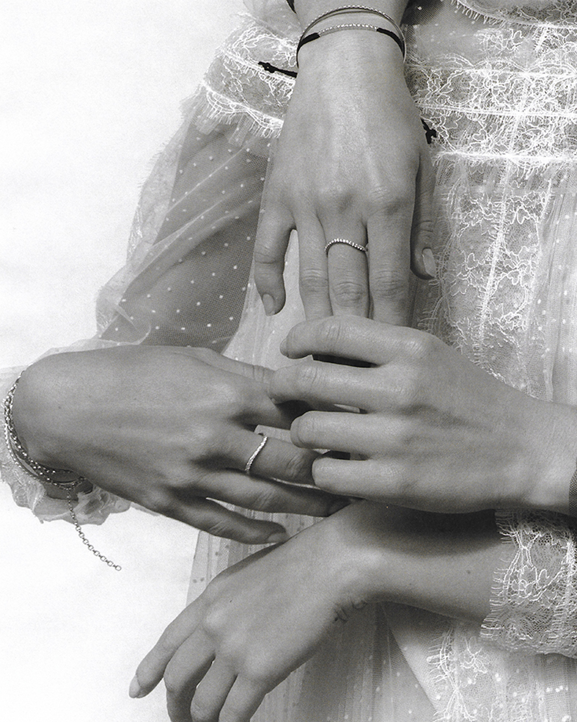 Close-up of two women's hands, adorned with minimalist rings and bracelets, resting gently against a white, semi-sheer, dotted dress.