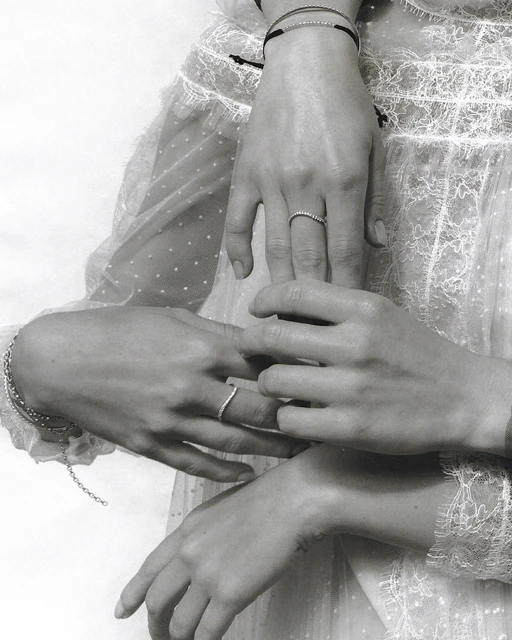 Close-up of two women's hands, adorned with minimalist rings and bracelets, resting gently against a white, semi-sheer, dotted dress.