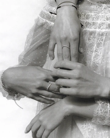 Close-up of two women's hands, adorned with minimalist rings and bracelets, resting gently against a white, semi-sheer, dotted dress.