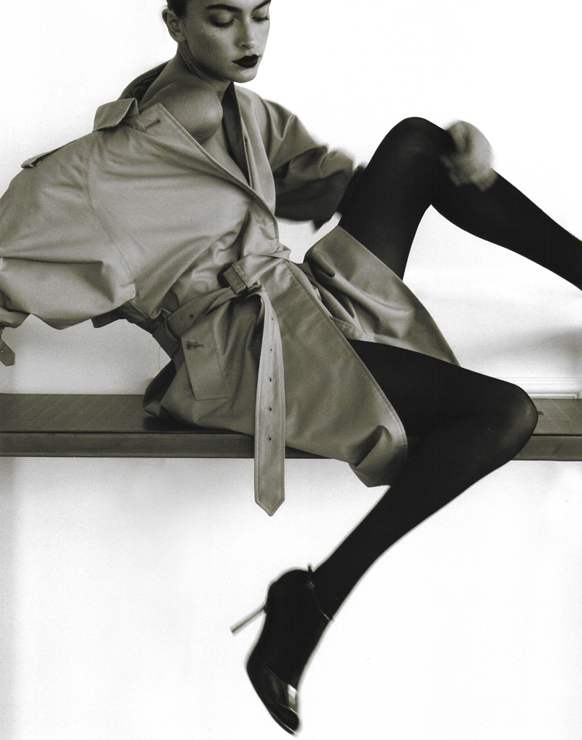 Model in oversized trench coat and black tights seated on a high bench, photographed by Adriana Roslin.