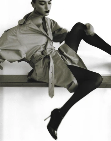 Model in oversized trench coat and black tights seated on a high bench, photographed by Adriana Roslin.