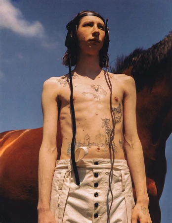 Shirtless young man with tattoos wearing headphones and white trousers standing against a clear blue sky