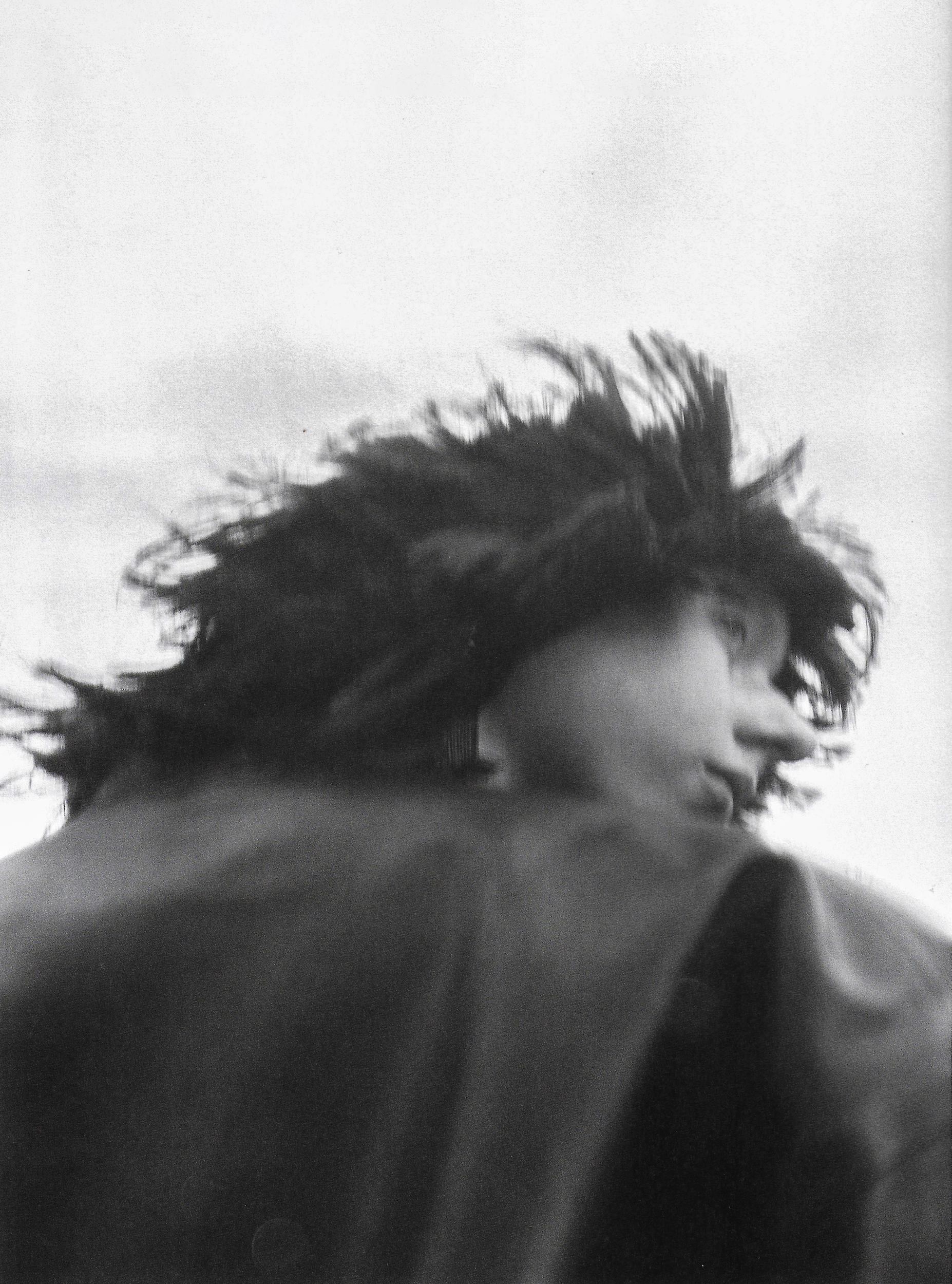 Monochrome portrait of a person with windblown dark hair, facing right, wearing a dark collared jacket against a bright sky.