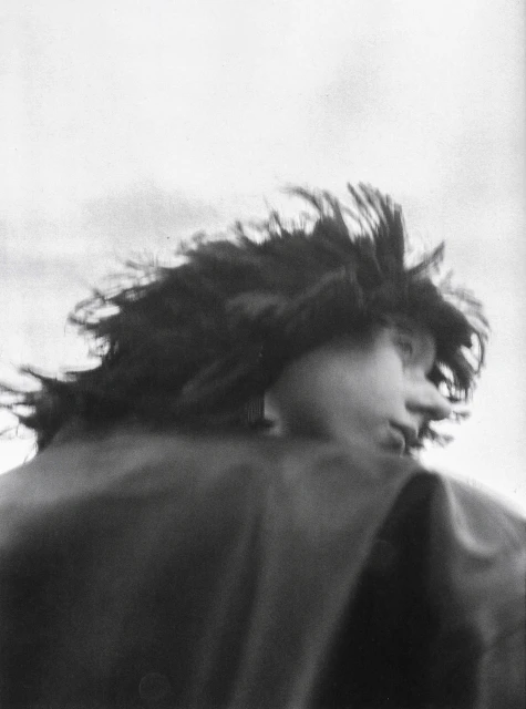 Monochrome portrait of a person with windblown dark hair, facing right, wearing a dark collared jacket against a bright sky.