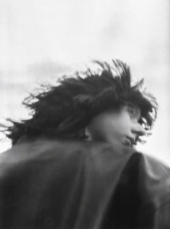 Monochrome portrait of a person with windblown dark hair, facing right, wearing a dark collared jacket against a bright sky.